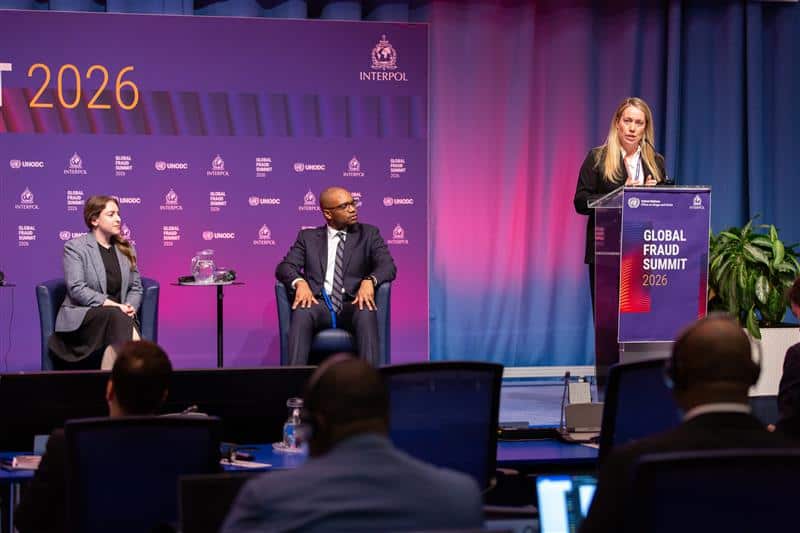 A photo of Stephanie Champion standing at a podium speaking at UNODC in front of a purple and pink background.